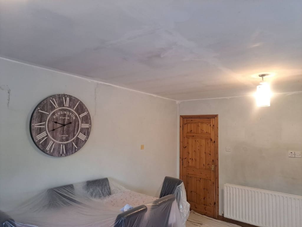 Dining room before decoration, with filler patches across the ceiling, pale walls, furniture wrapped in plastic and a large station clock.