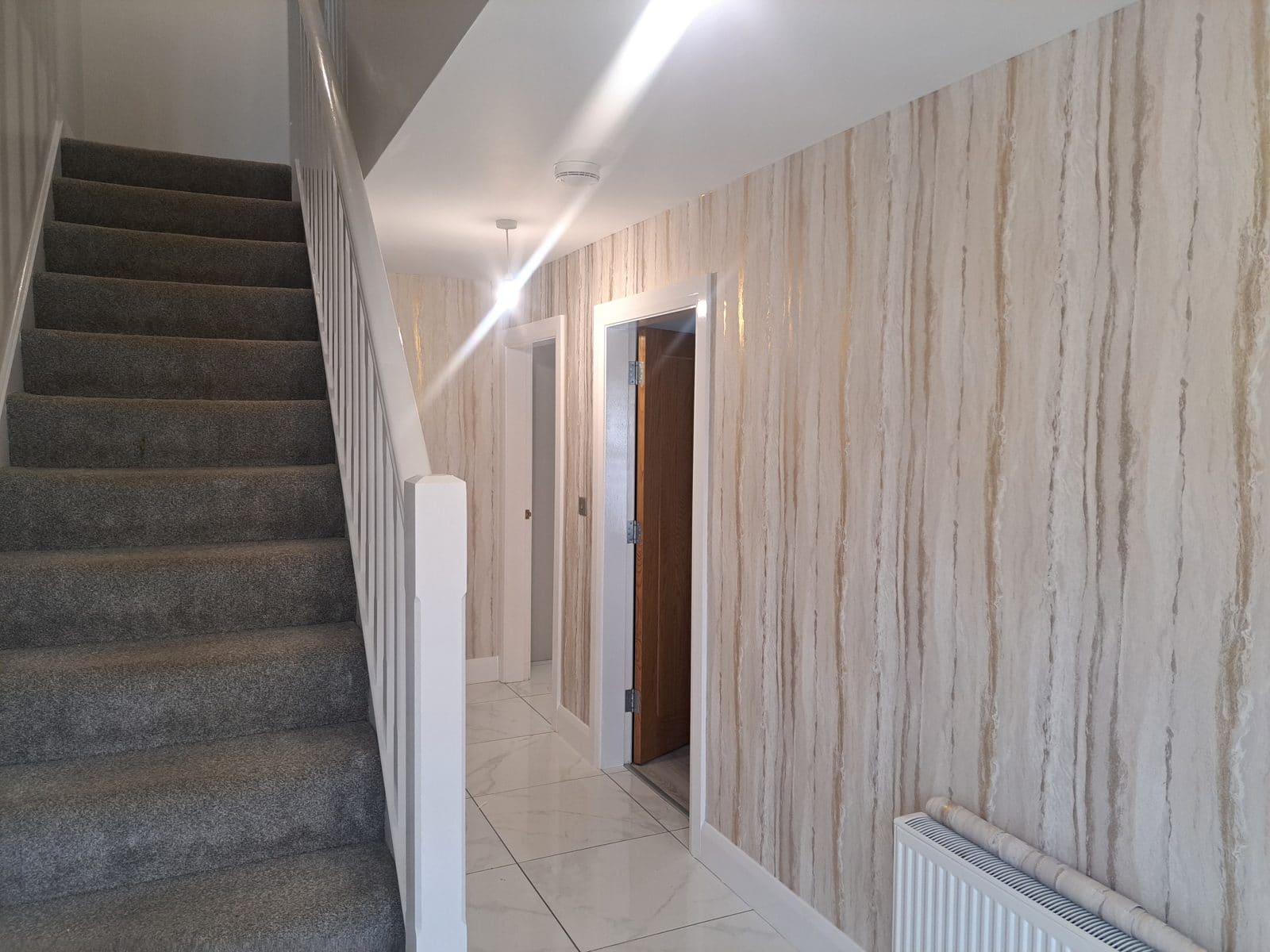 Hallway with a grey carpeted staircase and white spindles on the left, and a wall of beige and grey marble-effect wallpaper on the right. White marble-pattern floor tiles run down the hall.