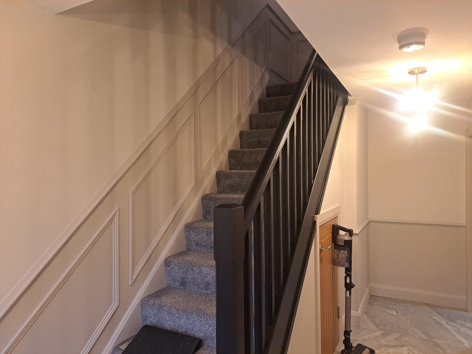 Staircase with cream raised-panel walls, a black handrail and black spindles, grey carpet, and a small wooden door under the stairs.